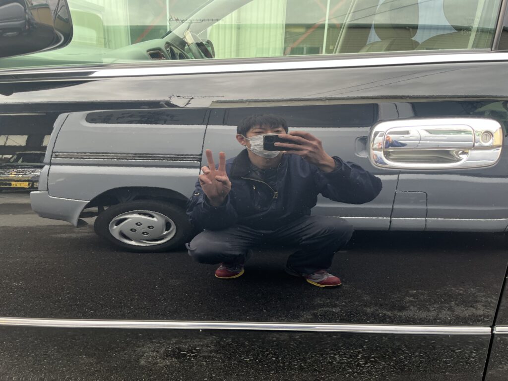 A mirror-like reflection on a polished black car, capturing the craftsman’s peace sign and expressing both craftsmanship and the joy of cars.
